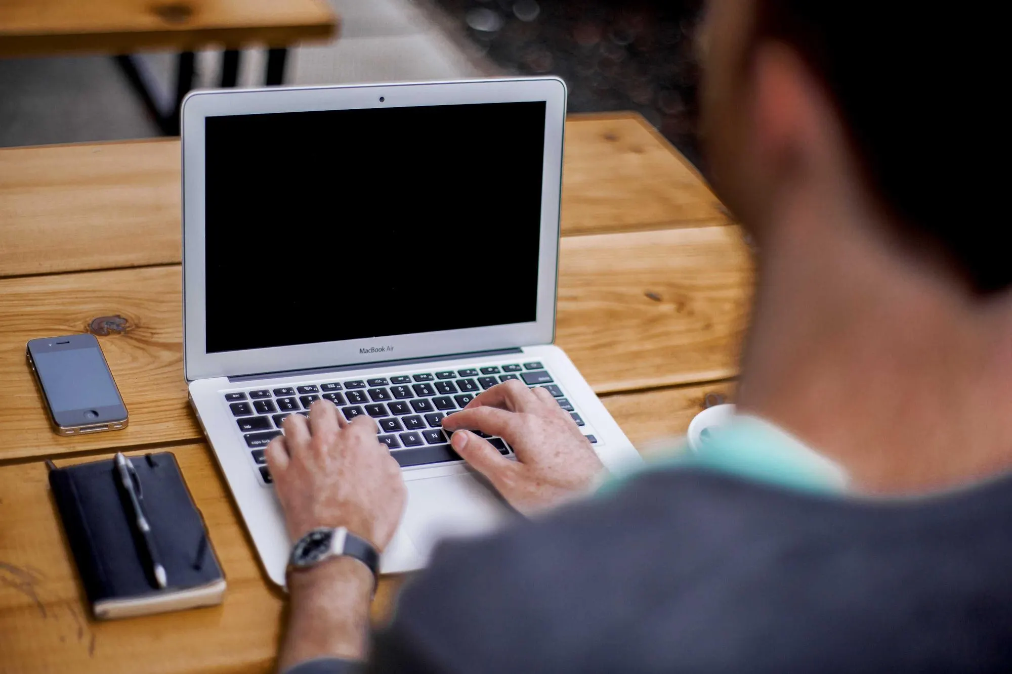 Person typing on a laptop at a wooden table, with a phone and notebook beside it